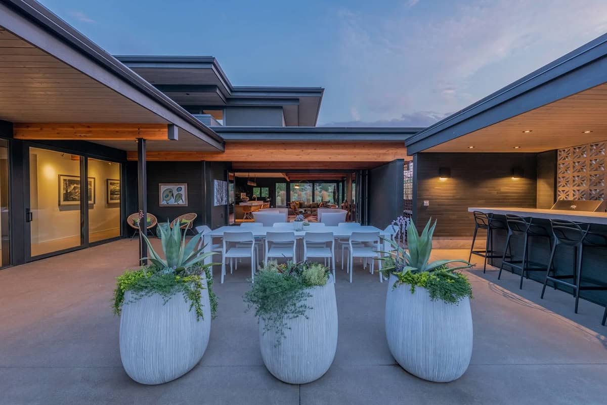 Outdoor patio at dusk with large ribbed white planters, white dining table, and view into illuminated interior