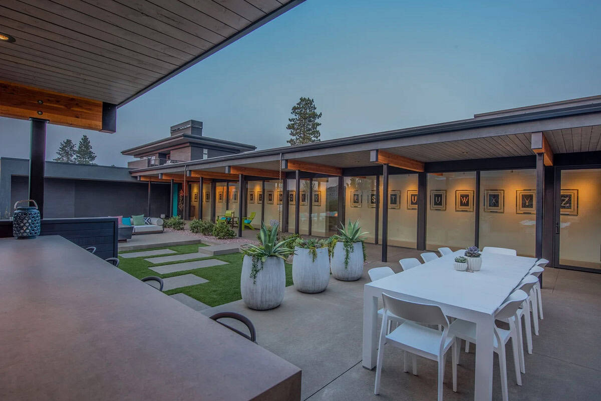 Covered outdoor dining area with white table and chairs, large planters, and floor-to-ceiling glazed gallery wall at dusk