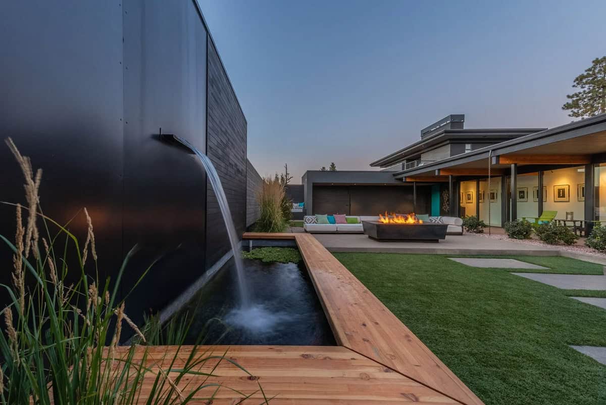 Backyard water feature with cascading waterfall into linear pool, cedar decking, and fire pit seating area at dusk
