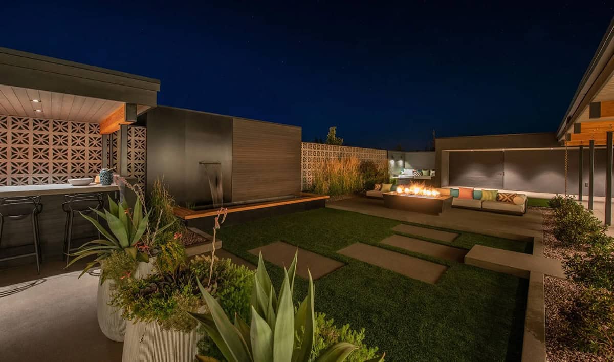 Nighttime backyard view with lit fire pit, outdoor sectional, ornamental grasses, decorative breeze block wall, and water feature
