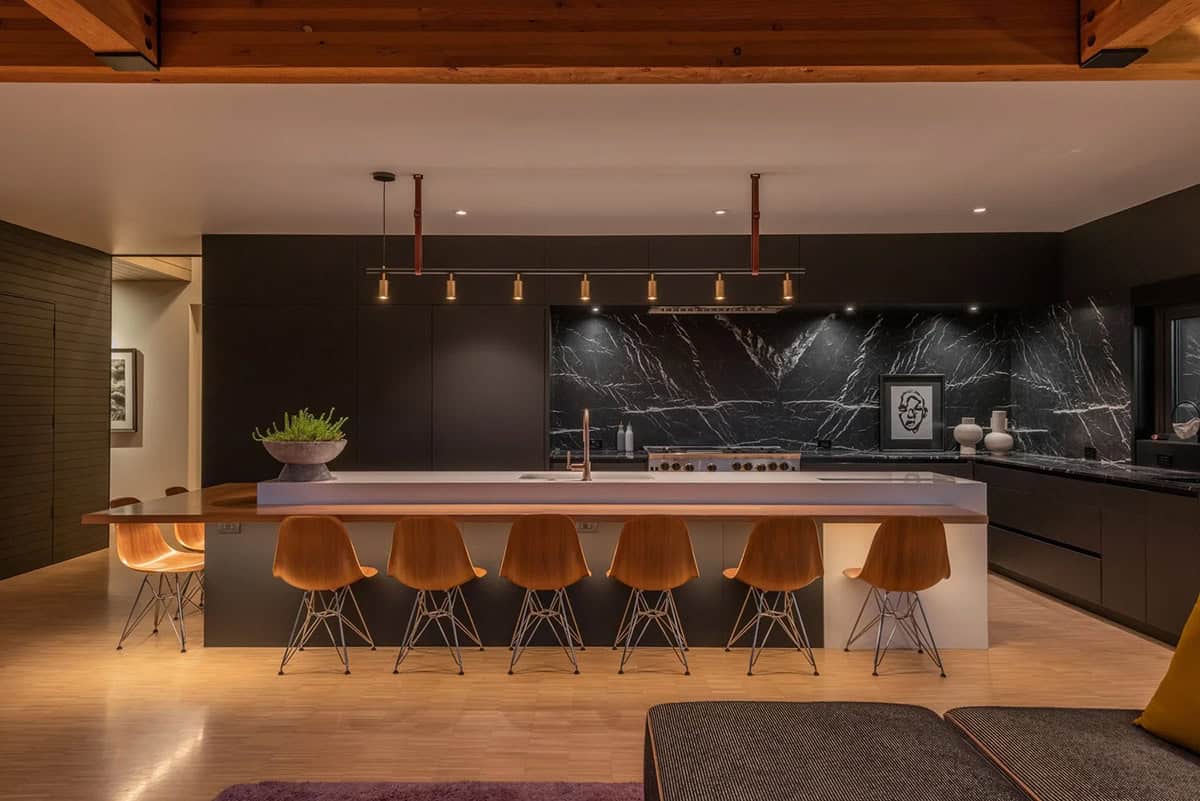 Evening kitchen interior with illuminated white island, six walnut bar stools, brass pendant lights, and black marble backsplash