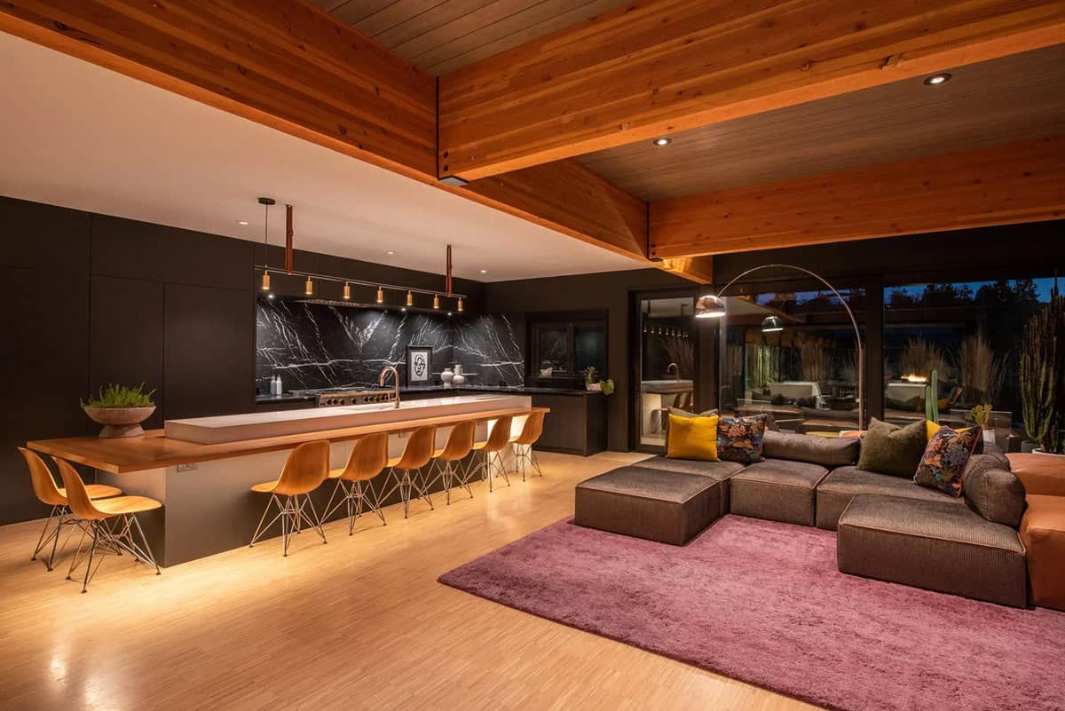 Open-plan kitchen and living area at night with wood beam ceiling, lit island, sectional sofa on purple rug, and courtyard view