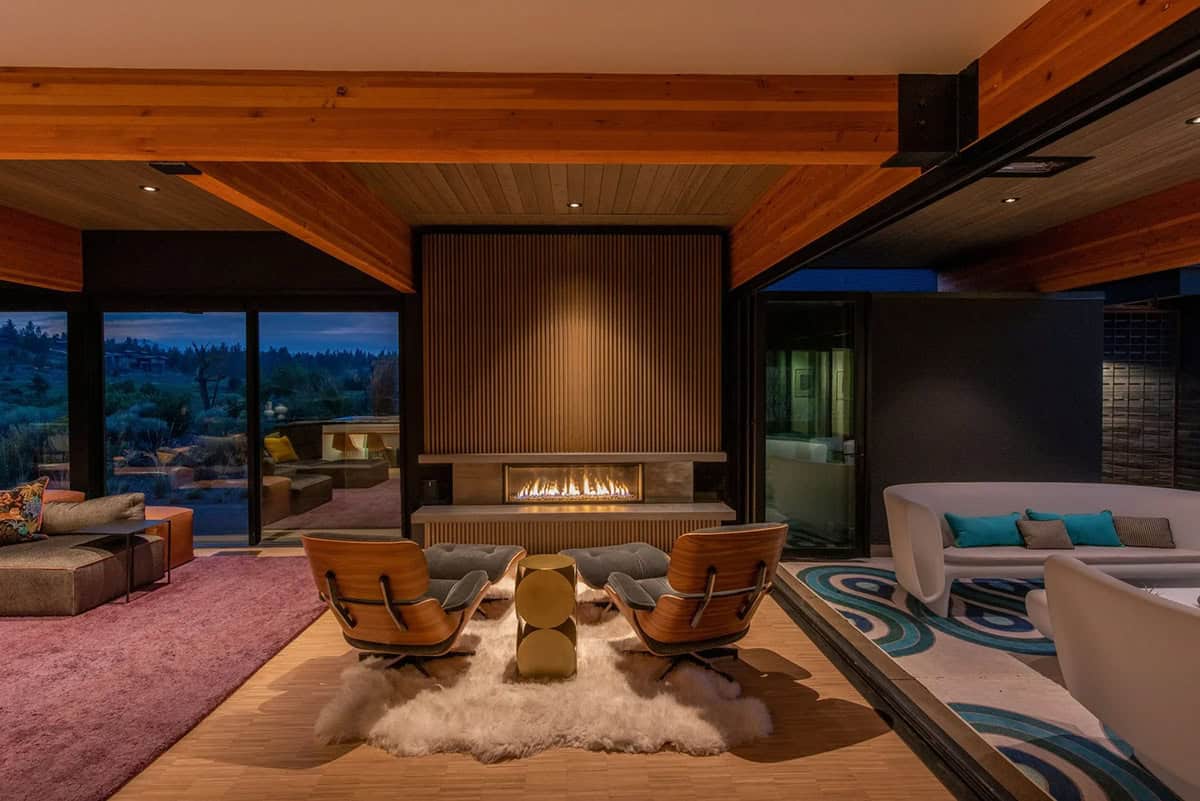 Evening living room with Eames lounge chairs on sheepskin rug, gold side table, lit linear fireplace, and slatted wood wall