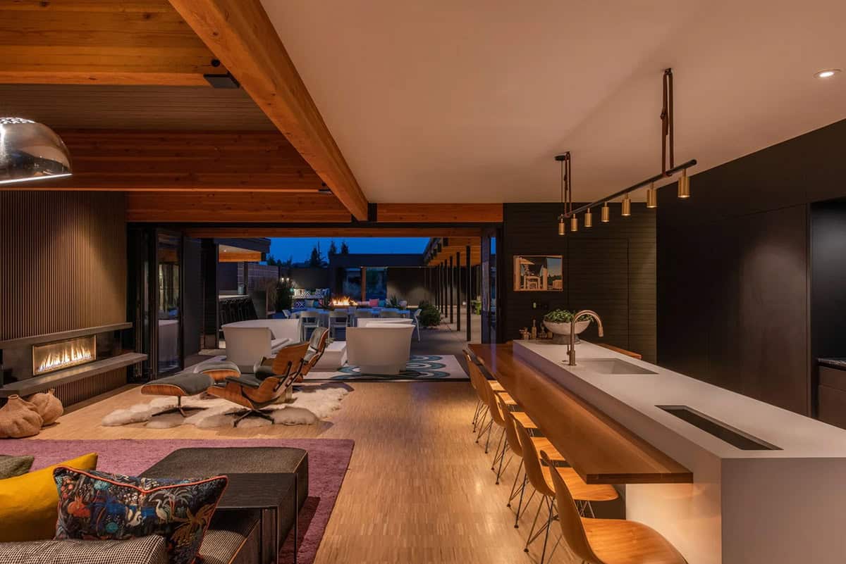Open-plan interior at dusk showing living area, kitchen island, fireplace, and fully open sliding glass walls to courtyard