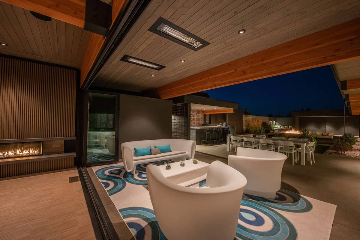 Covered outdoor lounge at night with white curved seating on swirl-patterned rug, ceiling heaters, linear fireplace, and outdoor bar beyond