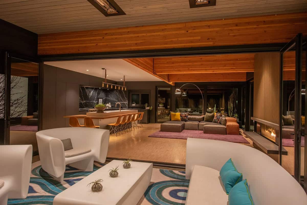 View from covered patio into open-plan interior at night showing white curved seating, kitchen island, and living area with wood beam ceiling
