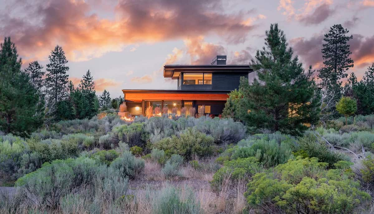 Dark modern home glowing at dusk among pine trees and sagebrush, with cedar accent band and large glazed openings