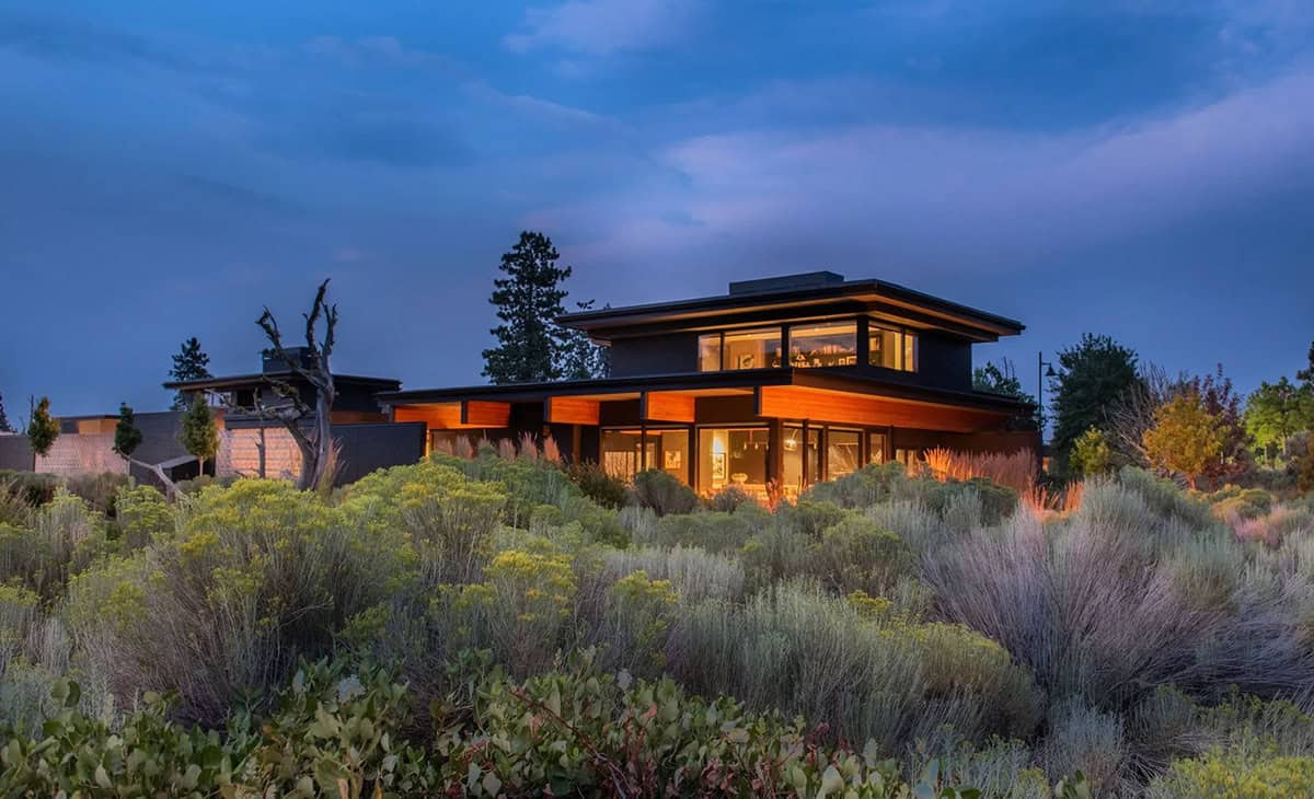 Illuminated two-story modern home at blue hour with cedar and dark stucco exterior, floor-to-ceiling windows, and native shrub landscape