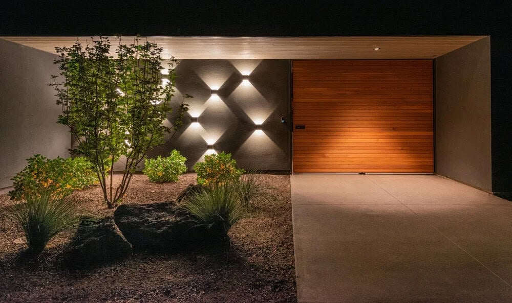 Night view of modern home entry with geometric wall sconces casting diamond light patterns beside a pivoting wood door