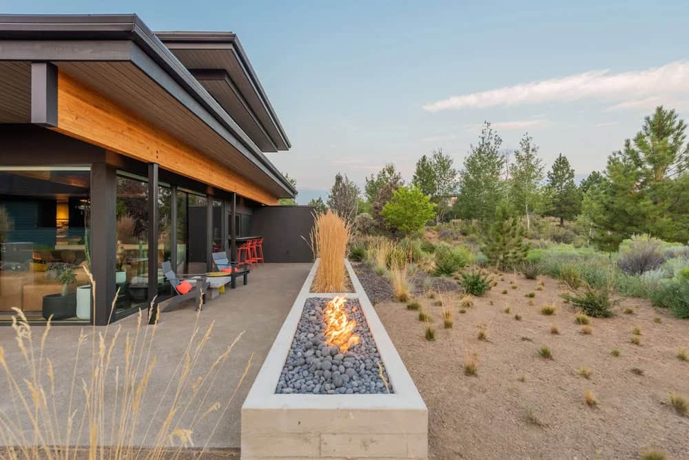 Linear concrete fire feature with river rock and open flame beside a dark modern home with cedar roofline and native landscape