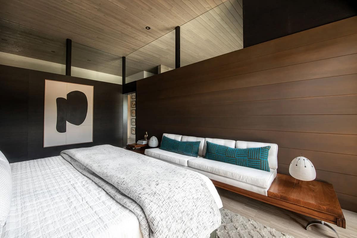 Primary bedroom with horizontal wood feature wall, white bench with teal pillows, abstract artwork, and wood plank ceiling