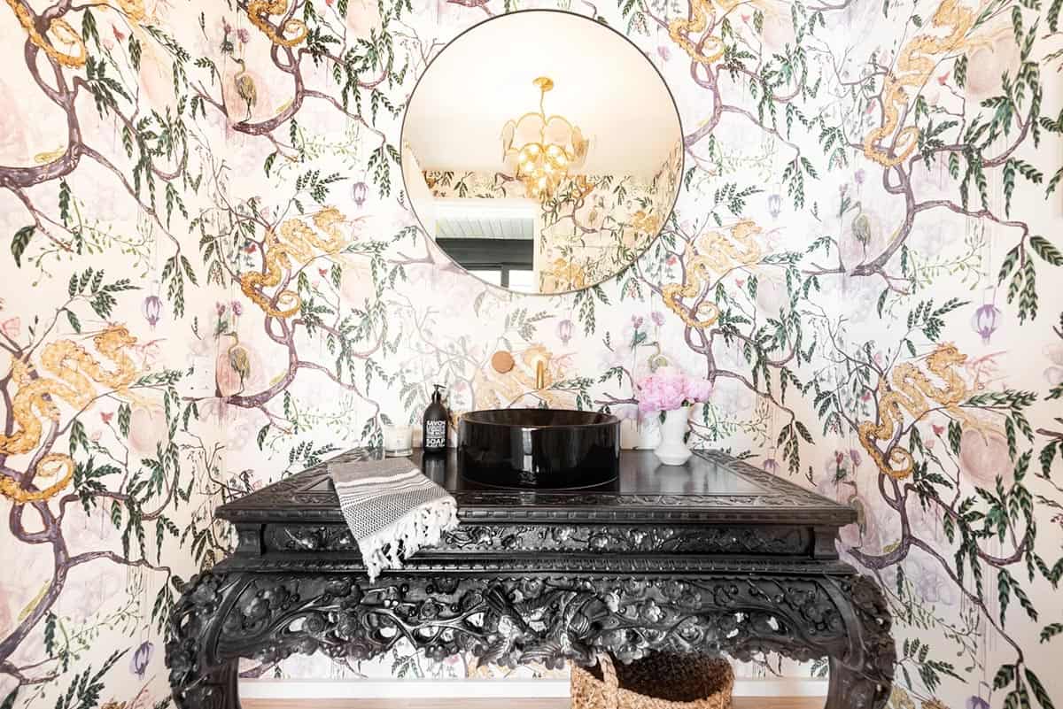 Powder room with dramatic botanical wallpaper, black carved vanity, round mirror, and gold chandelier reflection