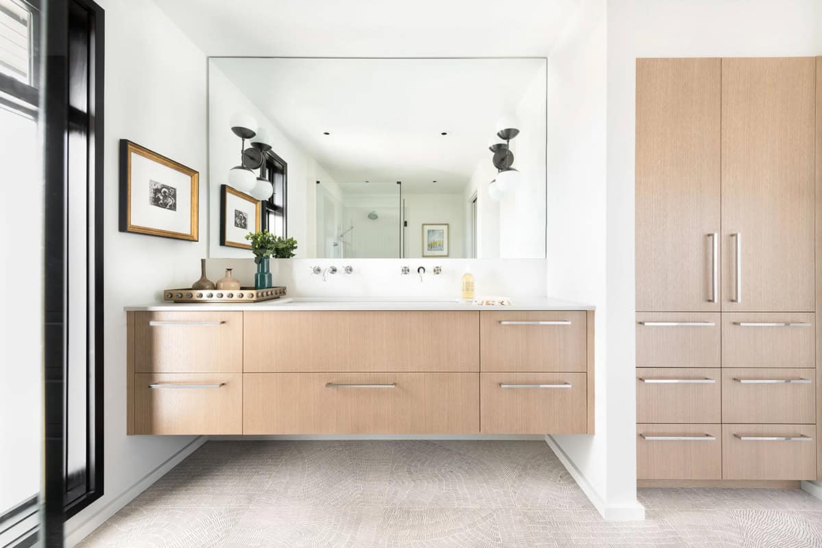Bright bathroom with floating light wood vanity, large frameless mirror, black globe sconces, textured floor tile, and framed artwork