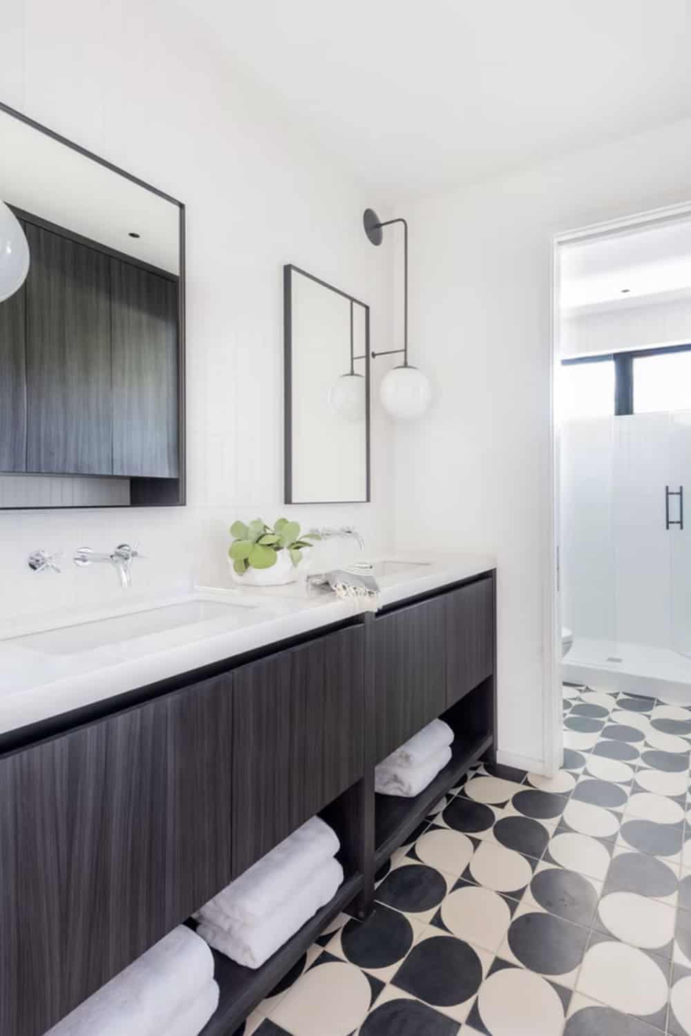 Modern guest bathroom with dark floating vanity, open towel storage, black globe sconce, bold geometric floor tile, and glass shower