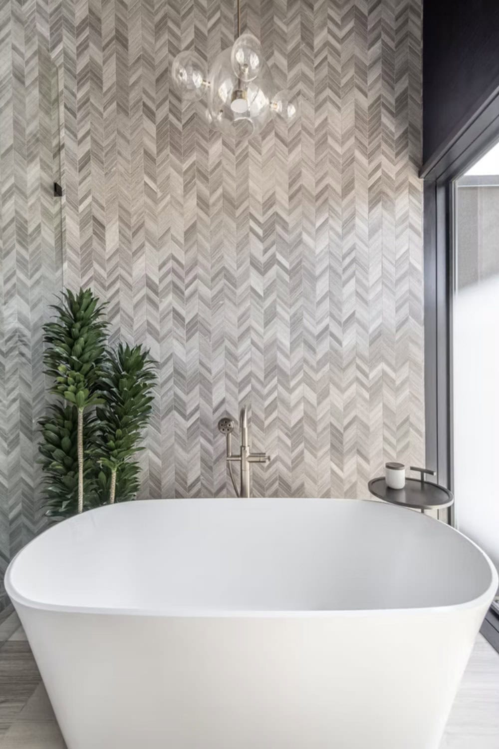 Freestanding white soaking tub against full-height chevron tile wall with bubble chandelier and tall indoor plant