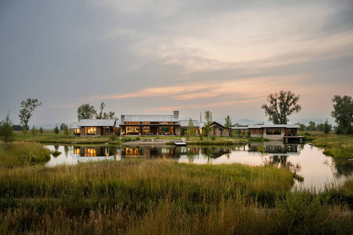 Wide view of a modern Montana ranch compound across a wetland pond at sunset