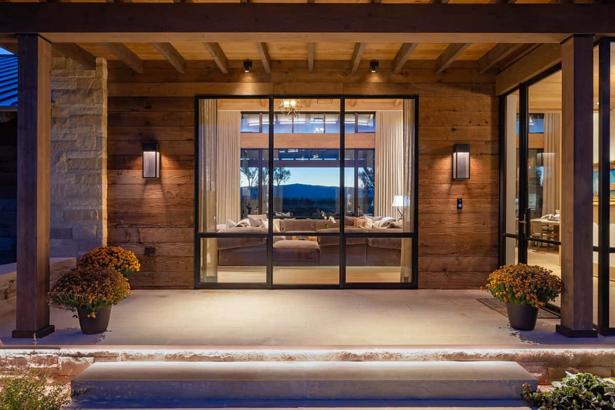 Covered entry porch with reclaimed wood siding, timber beams, and floor-to-ceiling glass doors at night
