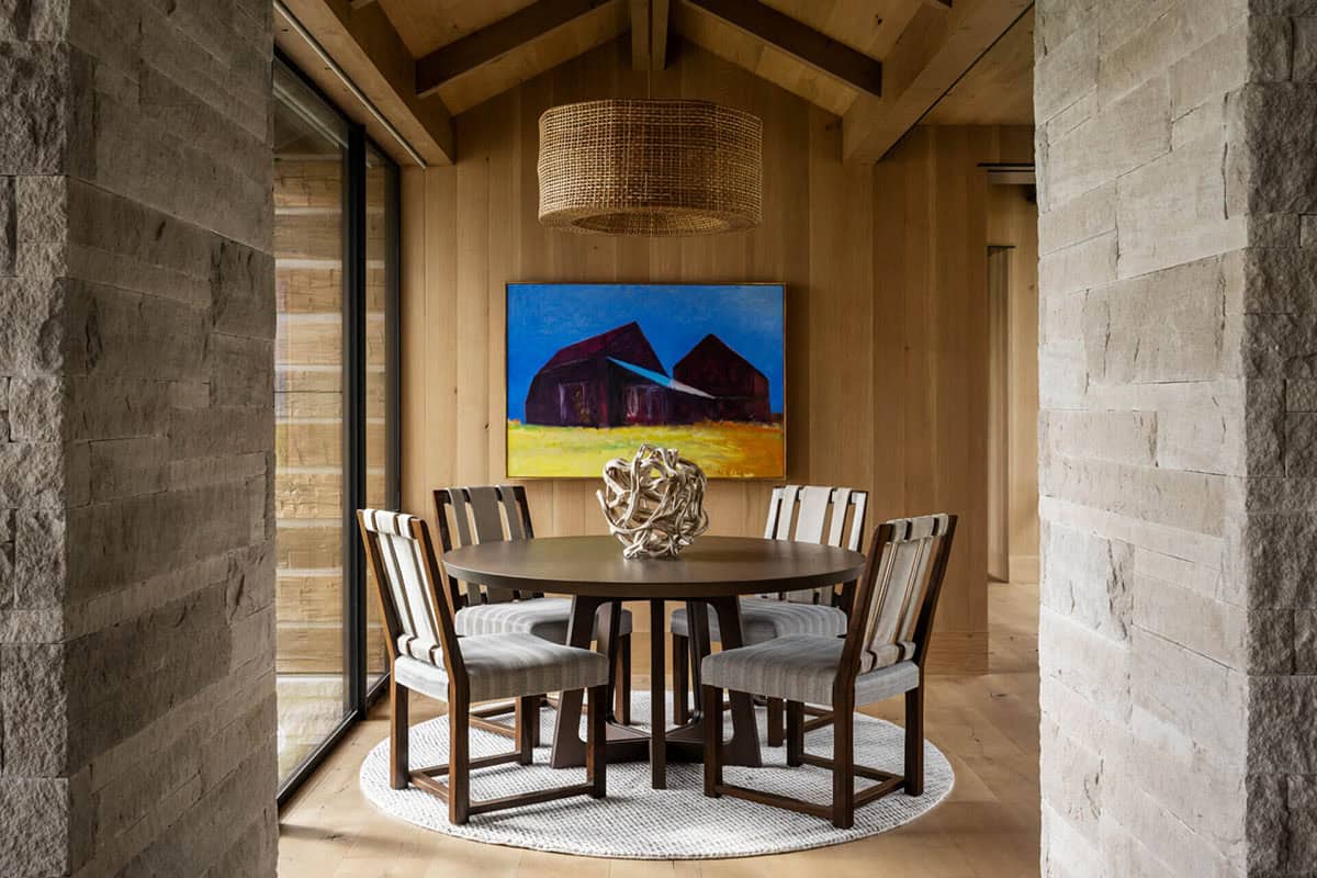 Intimate breakfast nook with round dark wood table, woven drum pendant, and barn painting on wood-paneled wall