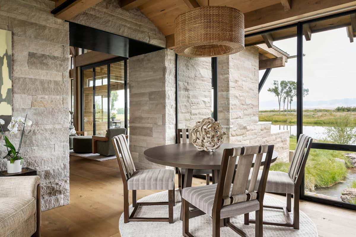 Round dining table with woven pendant light and floor-to-ceiling windows overlooking a pond and Montana grasslands