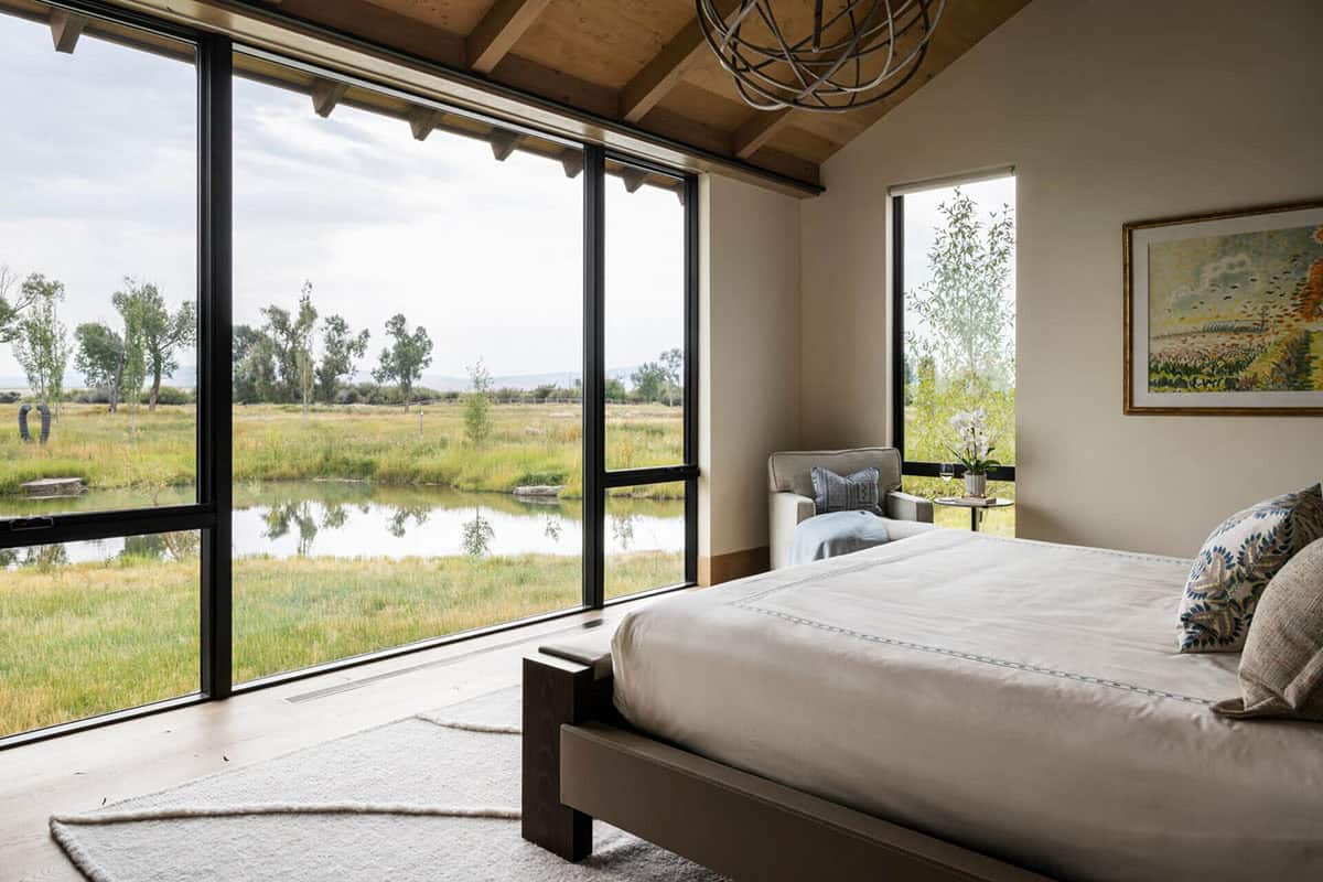 Primary bedroom with floor-to-ceiling black-framed windows overlooking a pond and native grasslands