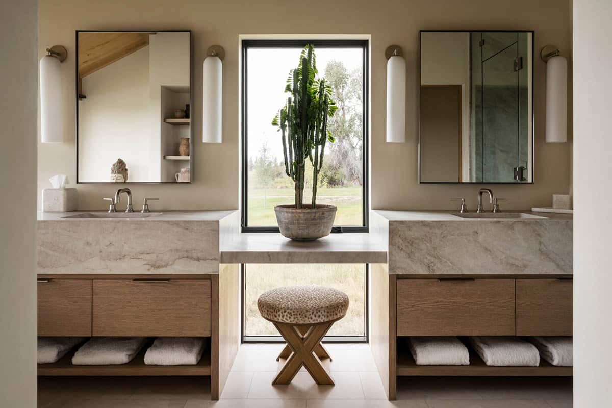 Primary bath with dual marble vanities, X-base stool, and window centered on a tall potted cactus