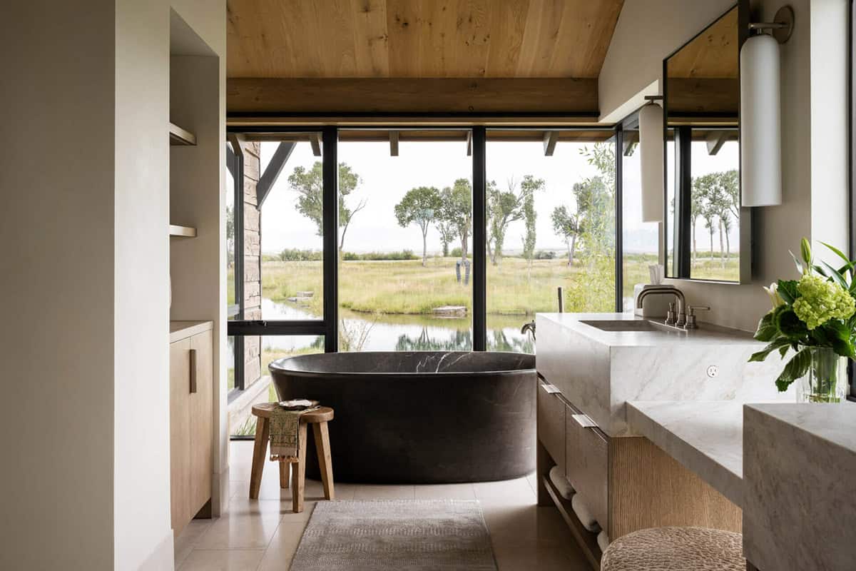 Freestanding black stone soaking tub facing panoramic windows with pond and grassland views