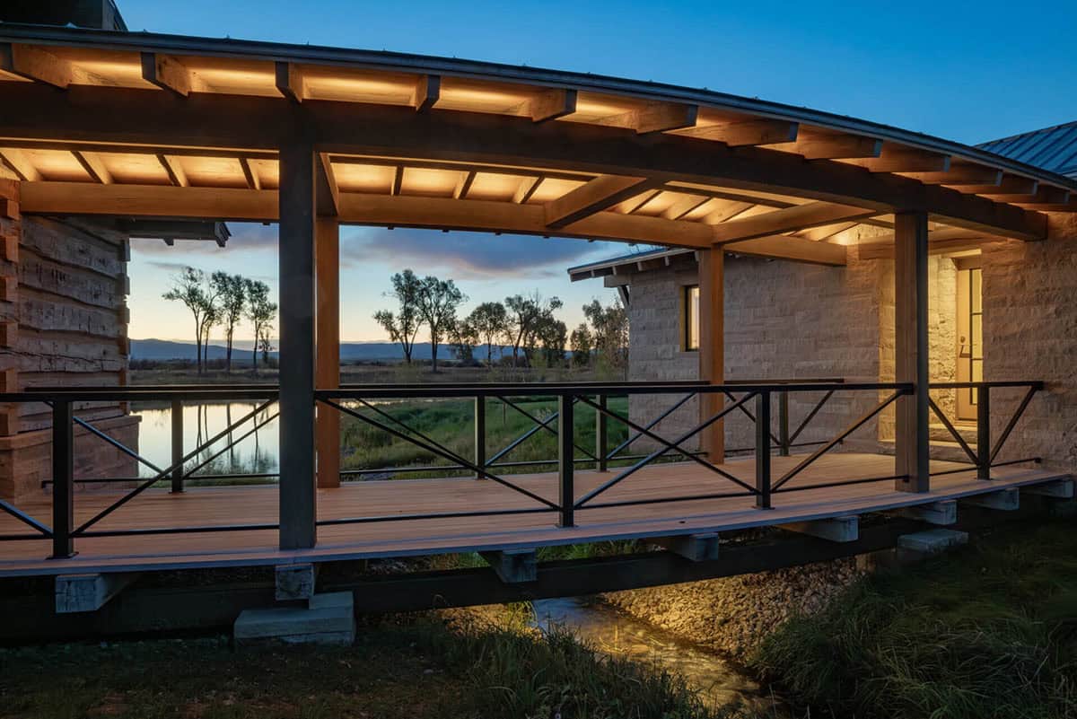 Lit wooden deck at dusk with steel cross-brace railing framing a pond and cottonwood tree reflection