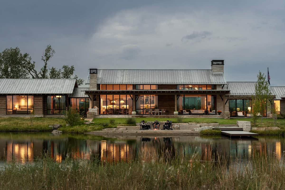 Family gathered around a fire pit at pond's edge at dusk, with the full ranch compound lit behind them