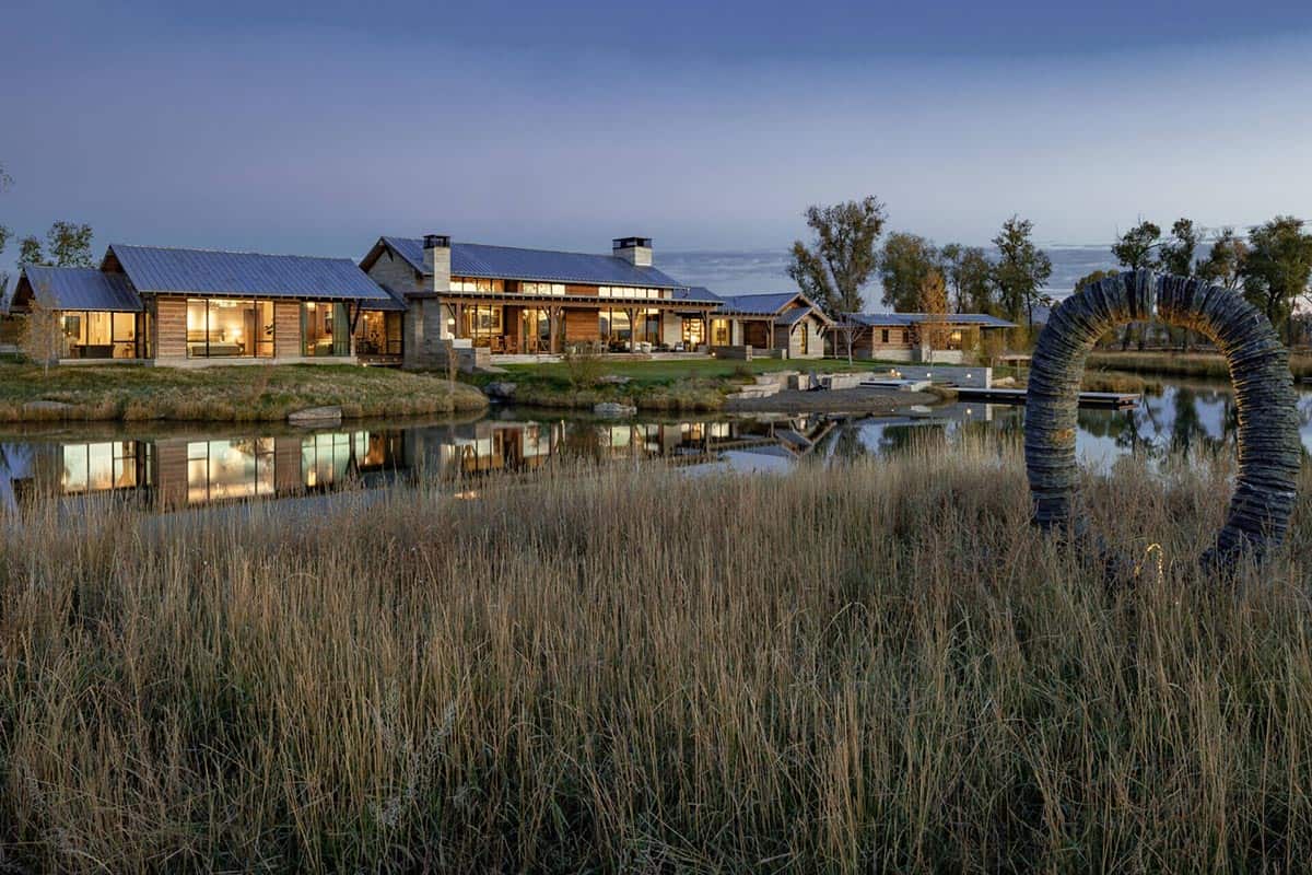 Montana ranch compound reflected in a pond at dusk, with dry autumn grasses and a curved sculpture in the foreground