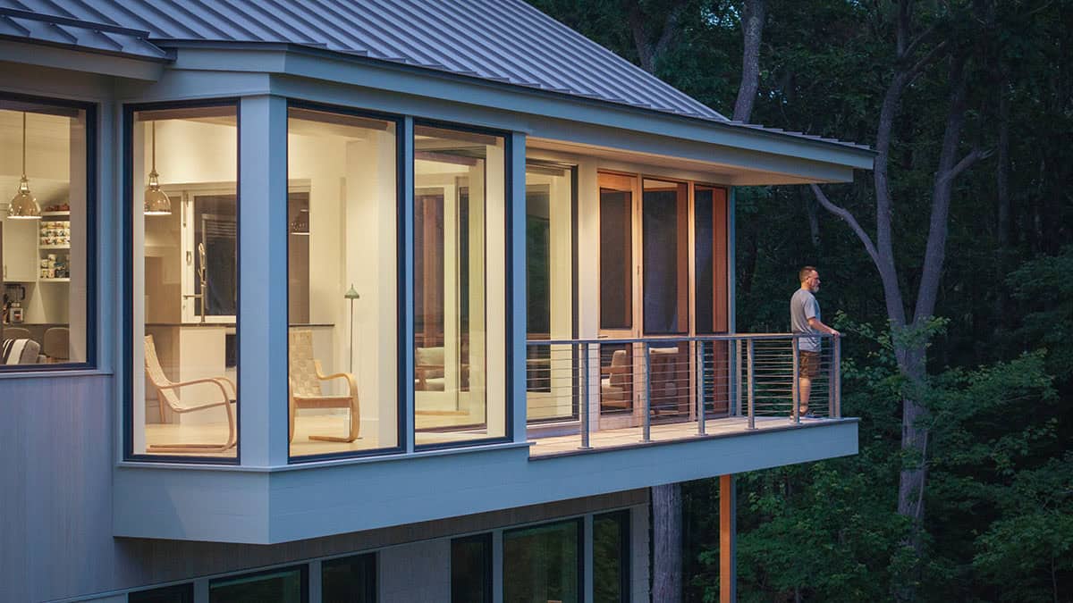 Man standing on upper-level balcony of modern treetop home at dusk, surrounded by forest canopy