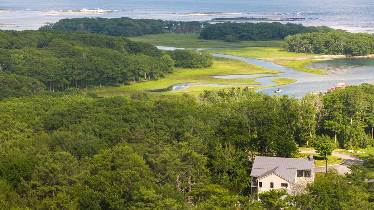 Aerial view of modern Maine home nestled among trees with coastal estuary and ocean beyond