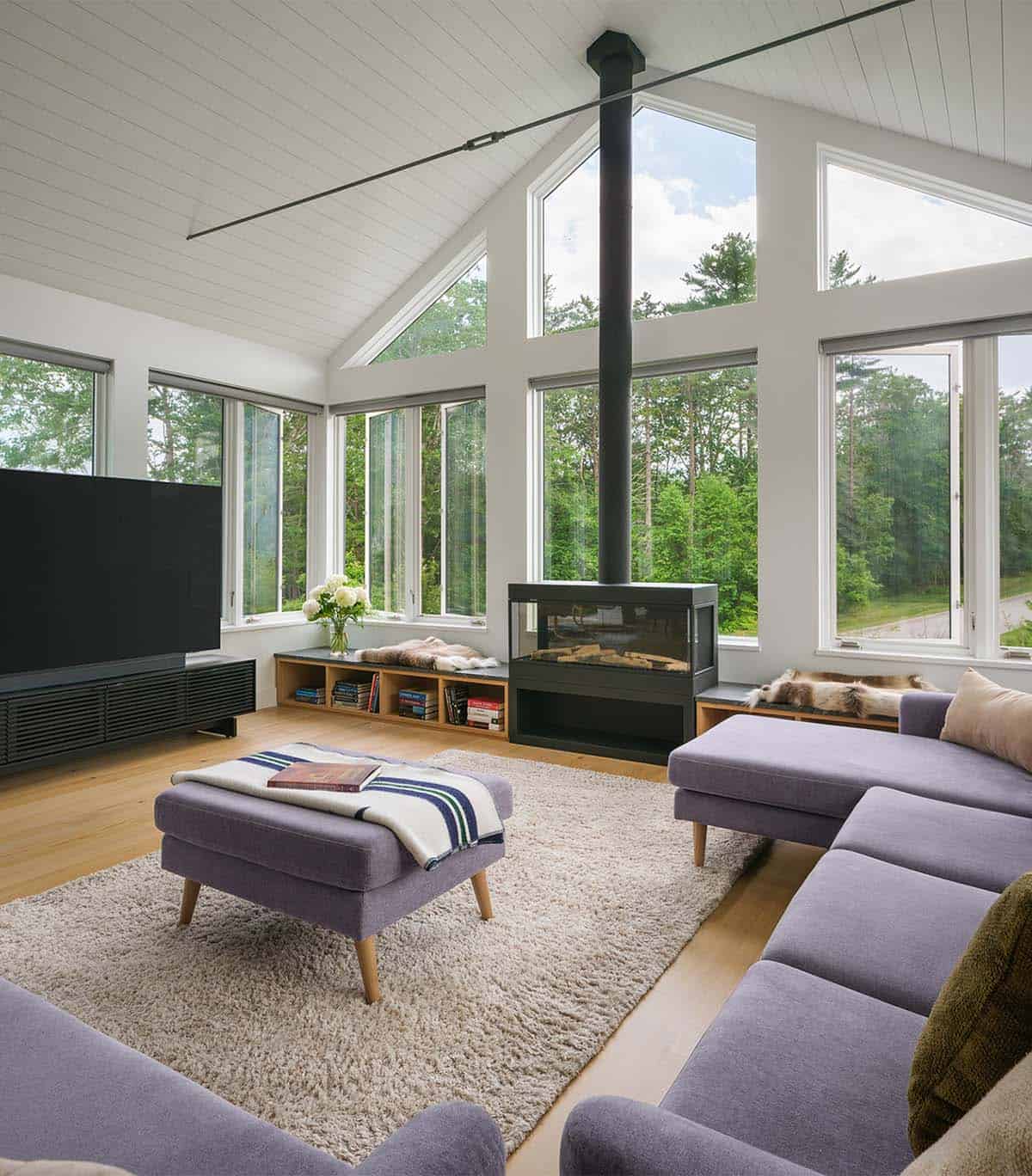 Vaulted living room with freestanding fireplace, shiplap ceiling, and wraparound forest windows