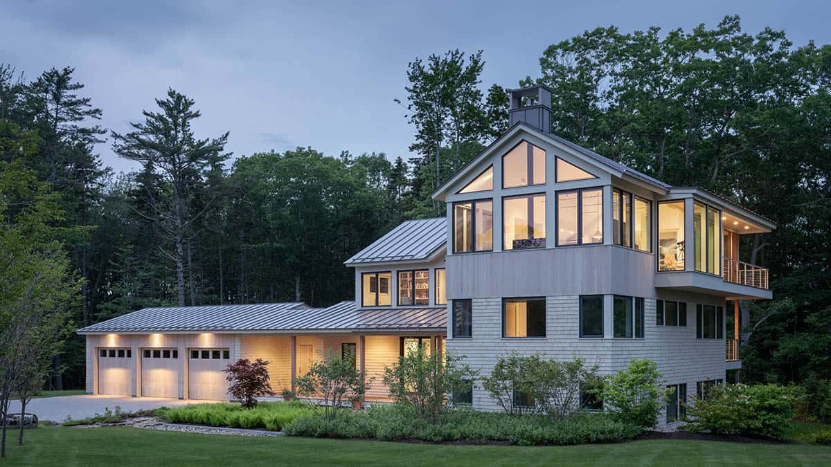Modern Maine home glowing at dusk with illuminated gable windows, garage wing, and landscaped entry