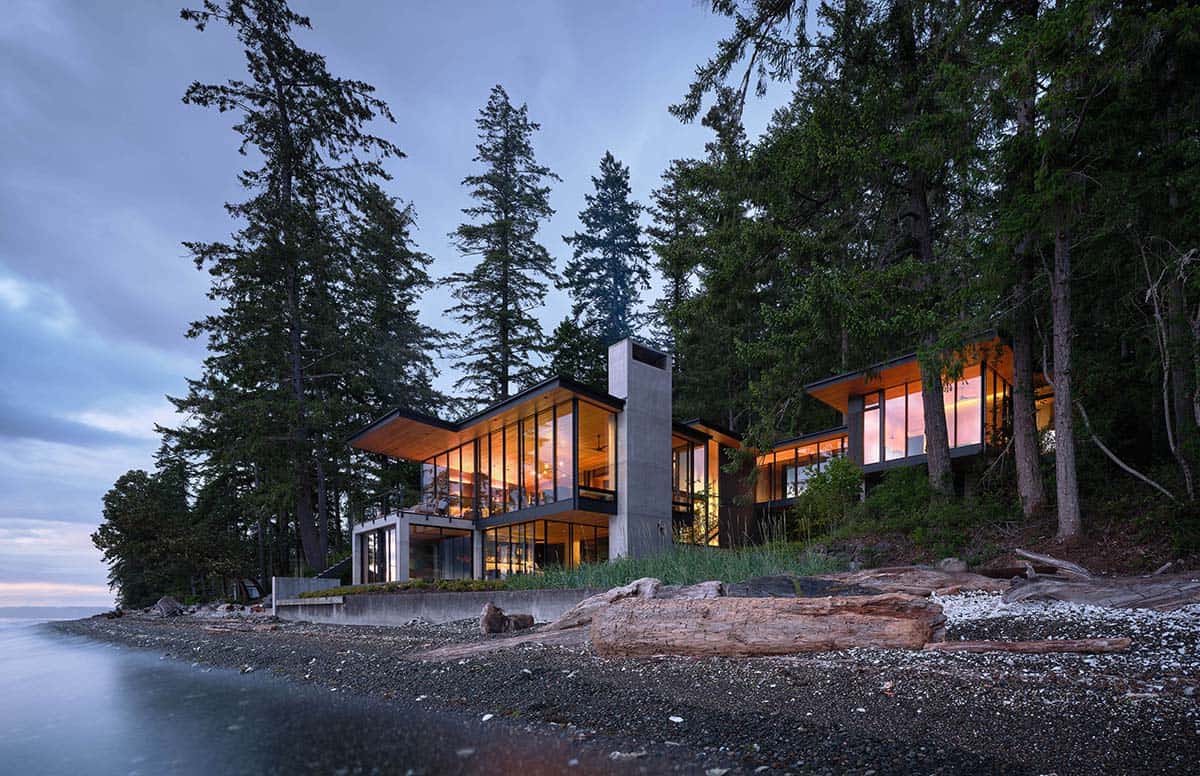 Modern waterfront home at dusk nestled among tall conifers on a rocky shoreline