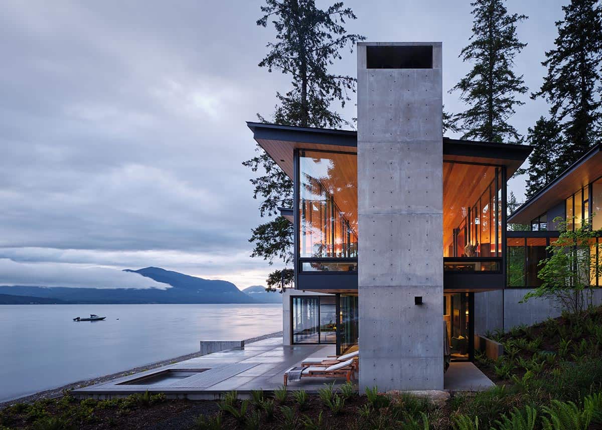Board-formed concrete chimney tower with floor-to-ceiling glazing overlooking a mountain lake at twilight