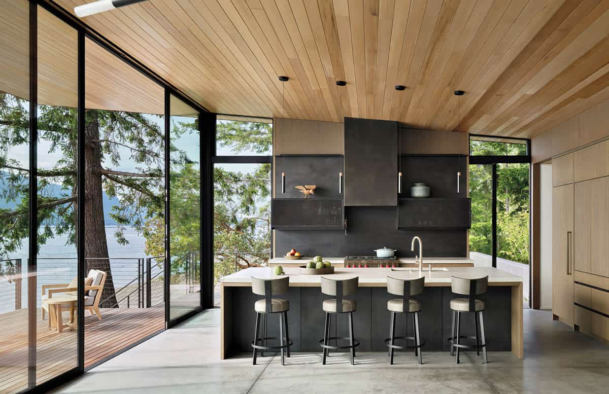 Modern waterfront kitchen with wood-plank ceiling, black cabinetry, light stone island with bar stools, and floor-to-ceiling windows overlooking a forested shoreline