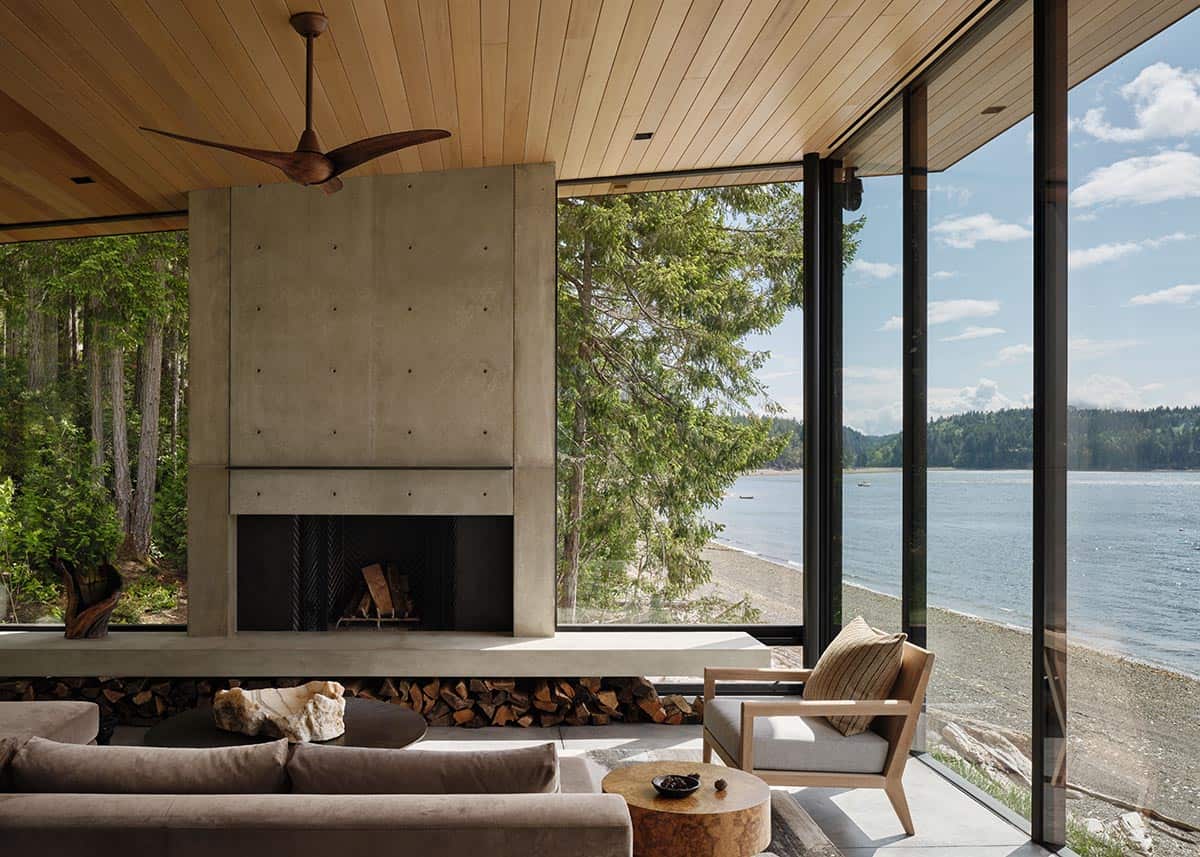 Living room with board-formed concrete fireplace, wood ceiling, and glass walls facing a pebbly shoreline