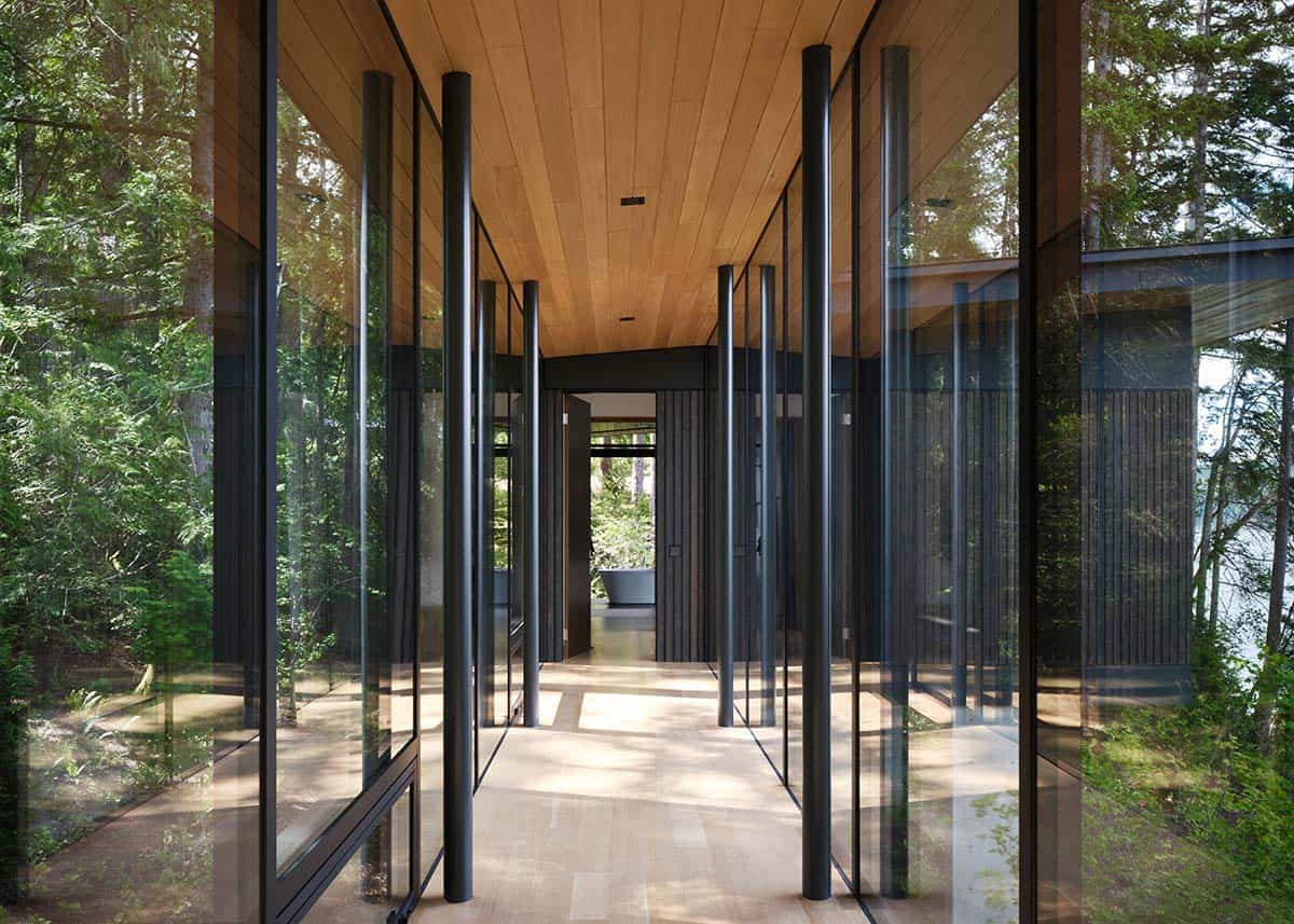 Glass-walled hallway corridor with black steel mullions reflecting surrounding forest