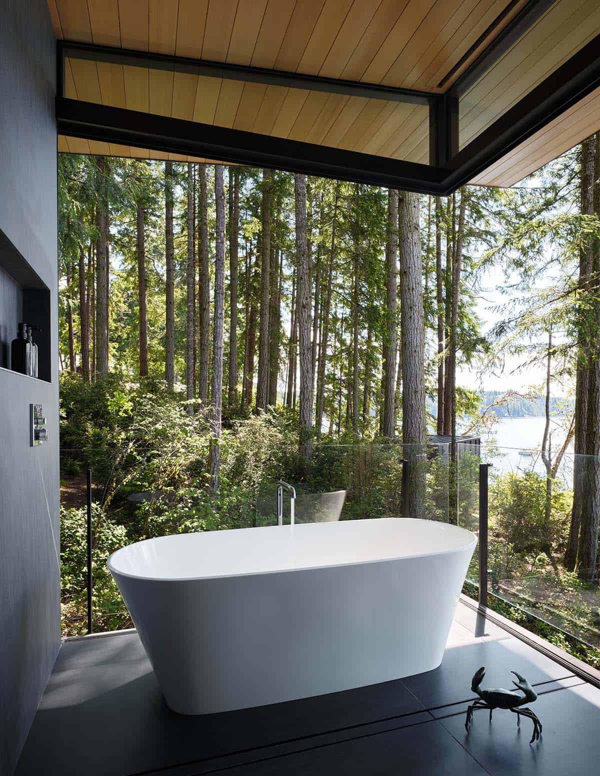 Freestanding white soaking tub at a glass corner with tall conifer forest and glimpse of lake