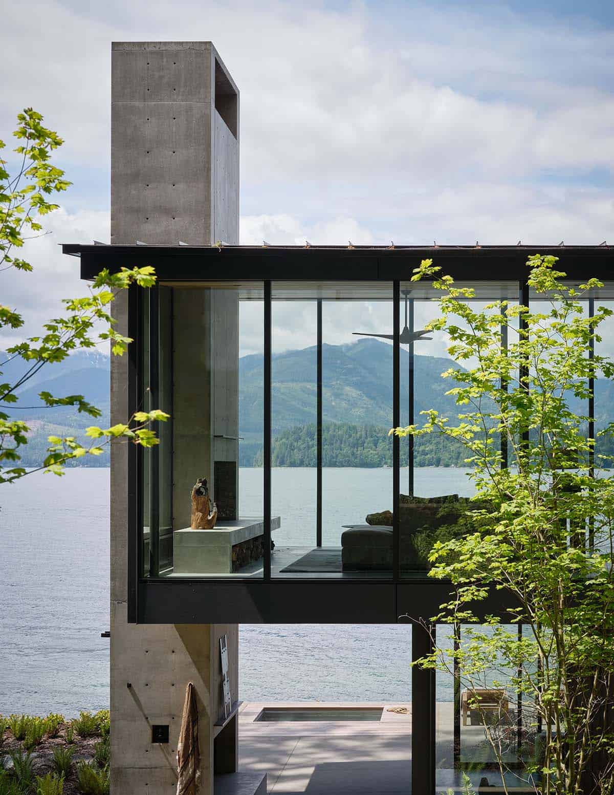 Glass living pavilion seen through spring maple foliage with lake and mountain backdrop