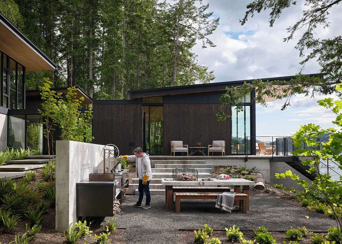 Person grilling at an outdoor kitchen beside a concrete dining table and dark wood-clad guest pavilion