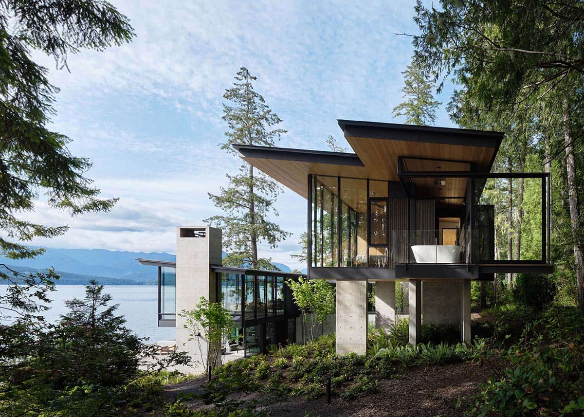 Cantilevered glass bedroom wing with flat cedar roof overhang rising above a forested hillside and lake