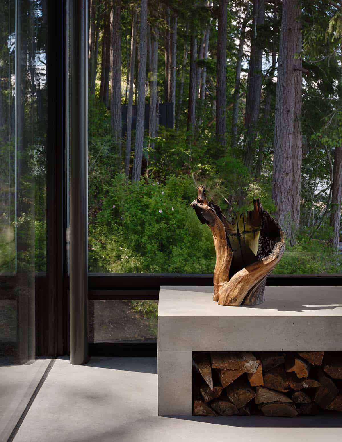 Driftwood sculpture on a concrete fireplace hearth with firewood storage and forest view through glass