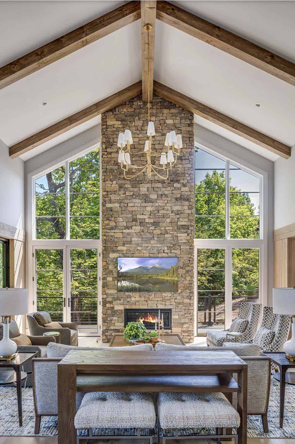 Soaring cathedral ceiling living room with stone fireplace flanked by tall windows overlooking forest