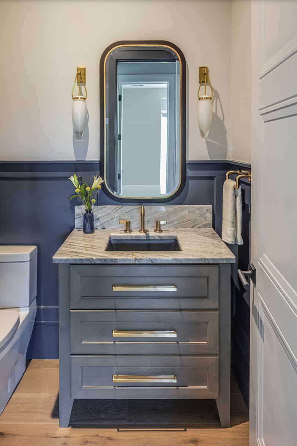 Powder room with slate-blue vanity, marble countertop, gold fixtures, and decorative mirror