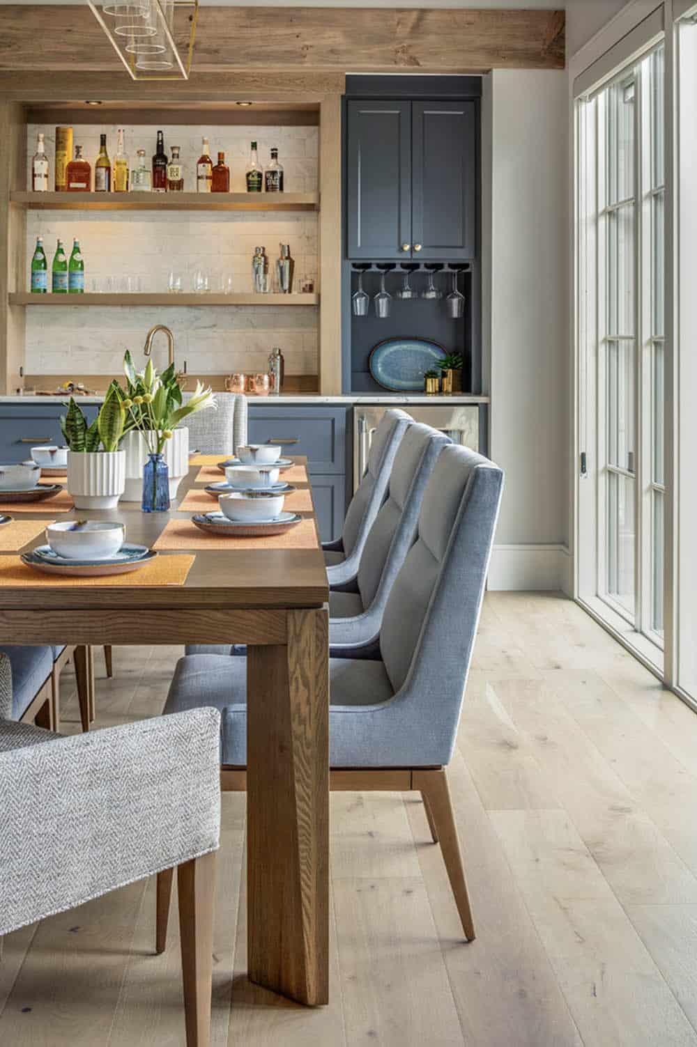 Dining table with blue upholstered chairs and built-in wet bar with open shelving and dark cabinetry