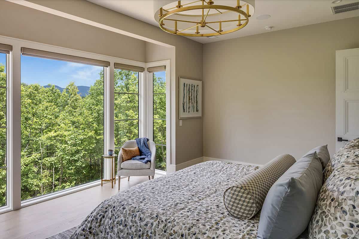 Primary bedroom with mountain and forest views through large windows, gold chandelier, and sitting chair