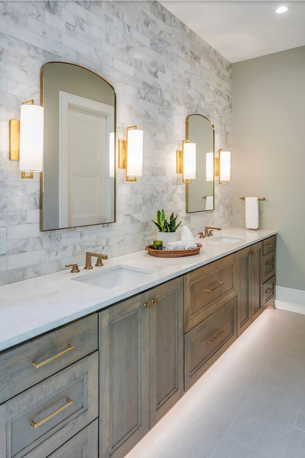 Primary bathroom double vanity with gray wood cabinets, marble tile wall, and gold arched mirrors