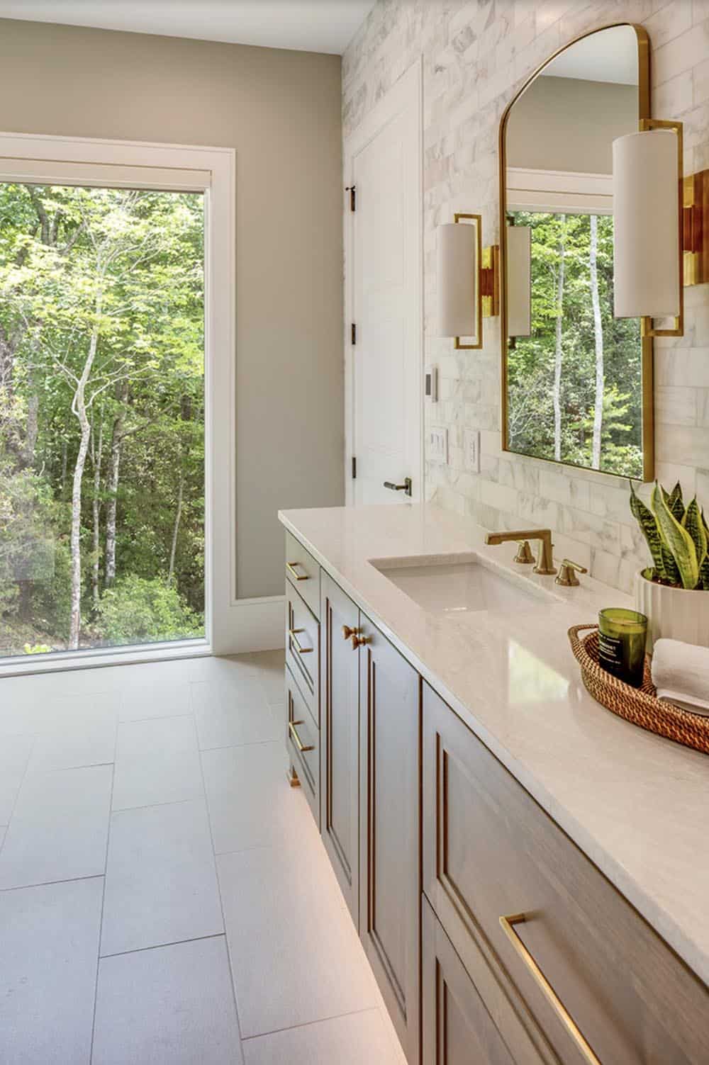 Primary bathroom single sink vanity with marble backsplash, gold fixtures, and forest window view