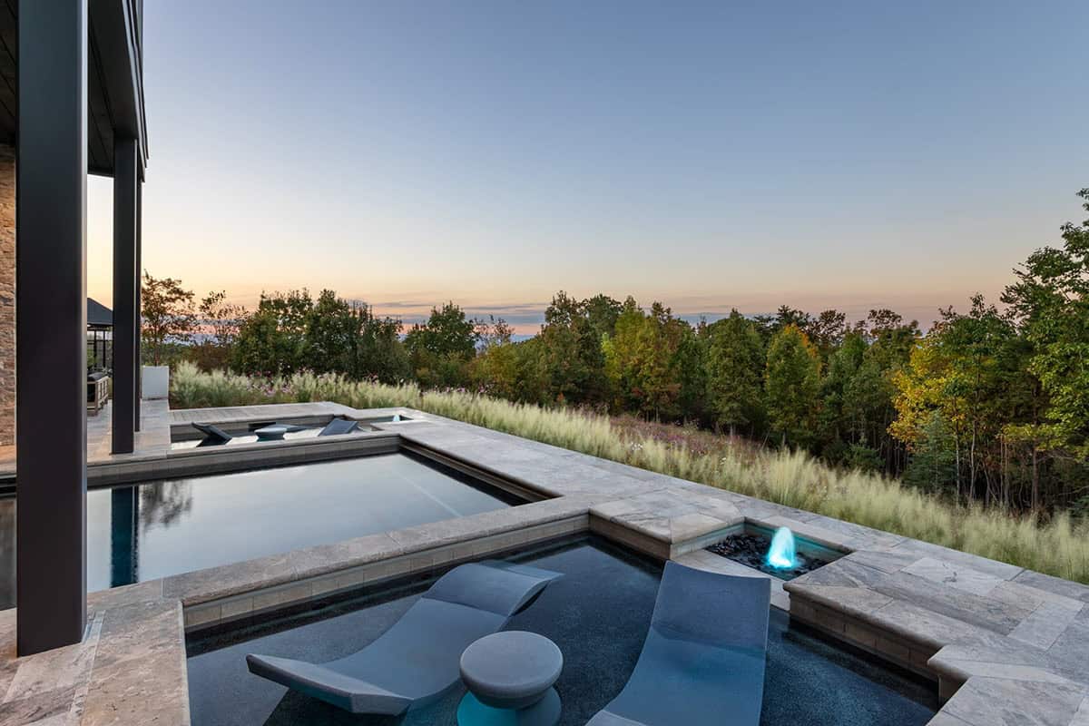 Infinity pool and spa with water feature overlooking forested hillside at sunset